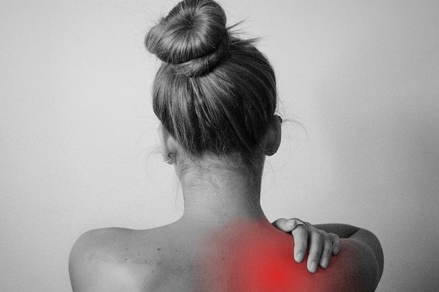 Frau mit hochgestecktem Haar, berührt sanft ihre schmerzhafte Schulter.